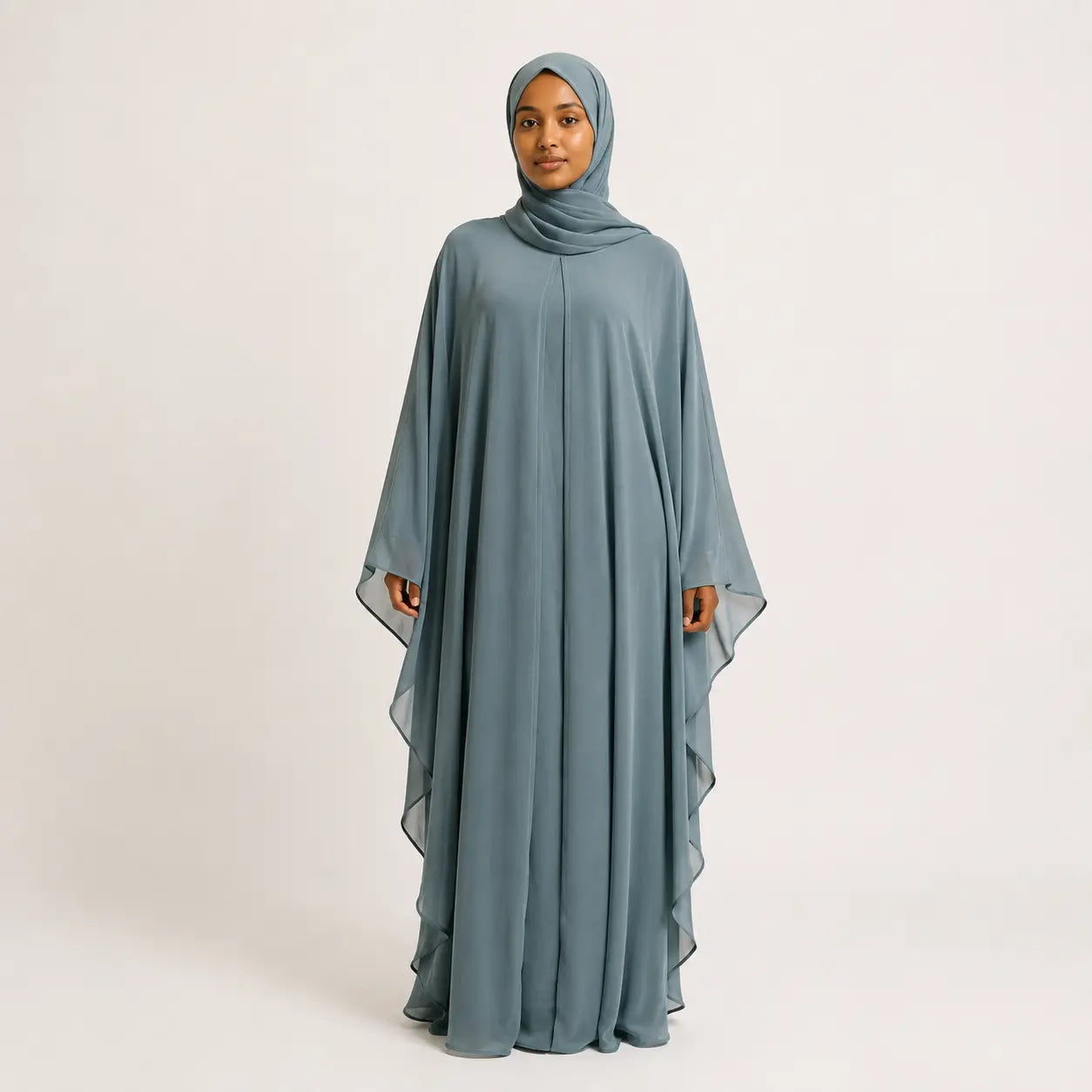 A woman wearing a flowing dusty blue-gray abaya with a matching hijab, standing against a plain light gray studio background.jade-green-chiffon-butterfly-farasha-abaya-hijab